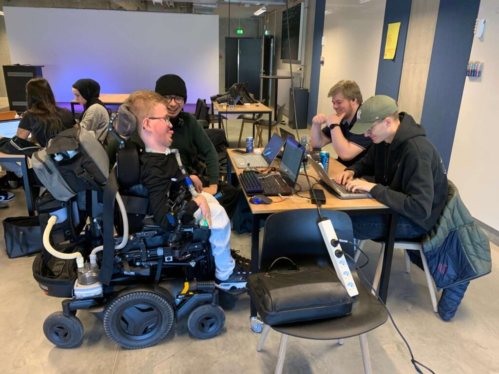 En gruppe mennesker sidder omkring et bord med bærbare computere og er engageret i diskussion. En person i en motoriseret kørestol er en del af gruppen. Lokalet har et moderne design med forskellige ting og udstyr synligt i baggrunden.