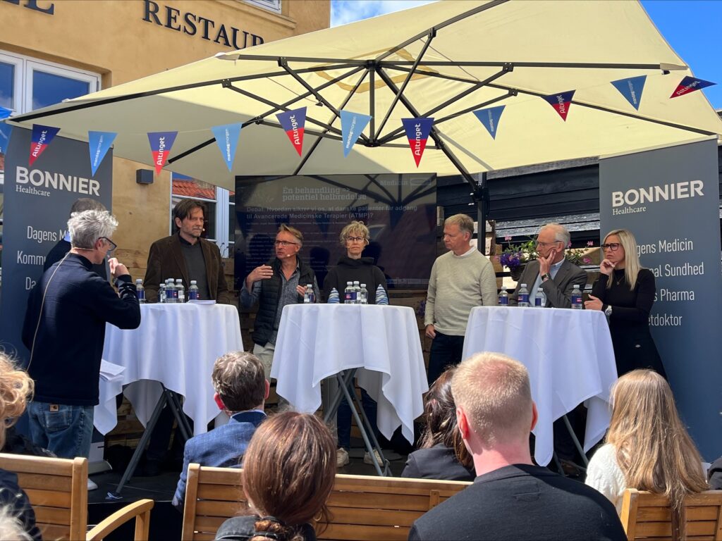 Et panel på seks personer står på en lille udendørs scene under en paraply og er engageret i diskussion. Bag dem er bannere med teksten 