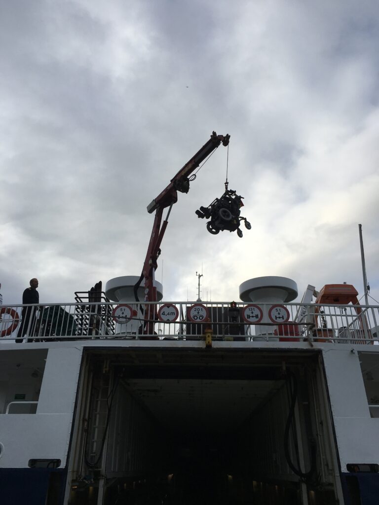 En kran på et skibsdæk sænker en stor motor- eller maskindel ned i fartøjets lastrum. Himlen er overskyet, og en person står nær kanten af dækket og observerer operationen. Ventilationsventilatorer og sikkerhedsudstyr er synlige på øverste dæk.