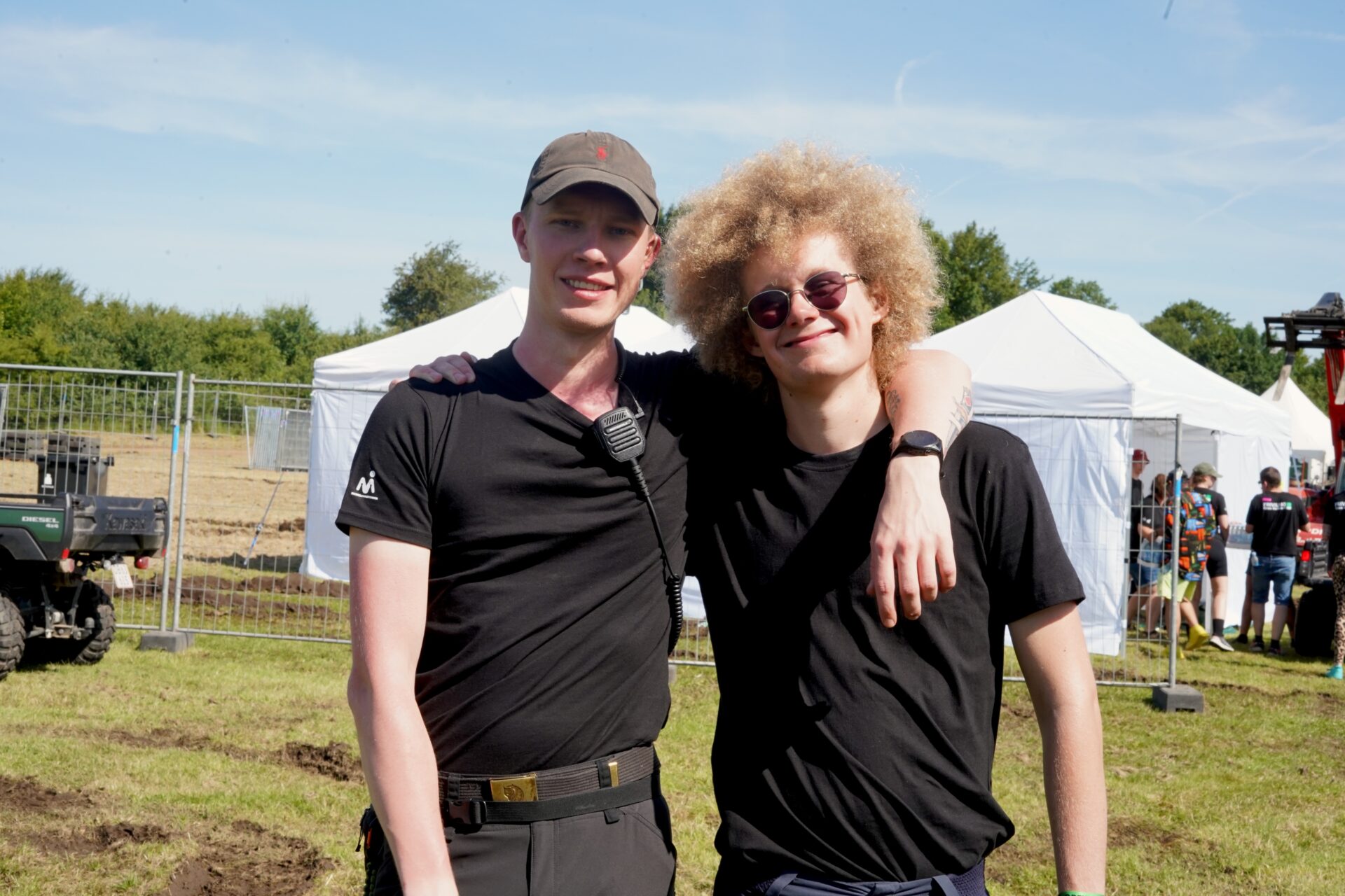 To unge mænd står udendørs foran hvide telte og metalhegn. Begge smiler, mens de poserer sammen, med den ene mand iført kasket og sort skjorte, og den anden iført solbriller og en sort t-shirt. Himlen ovenover er klar og blå.