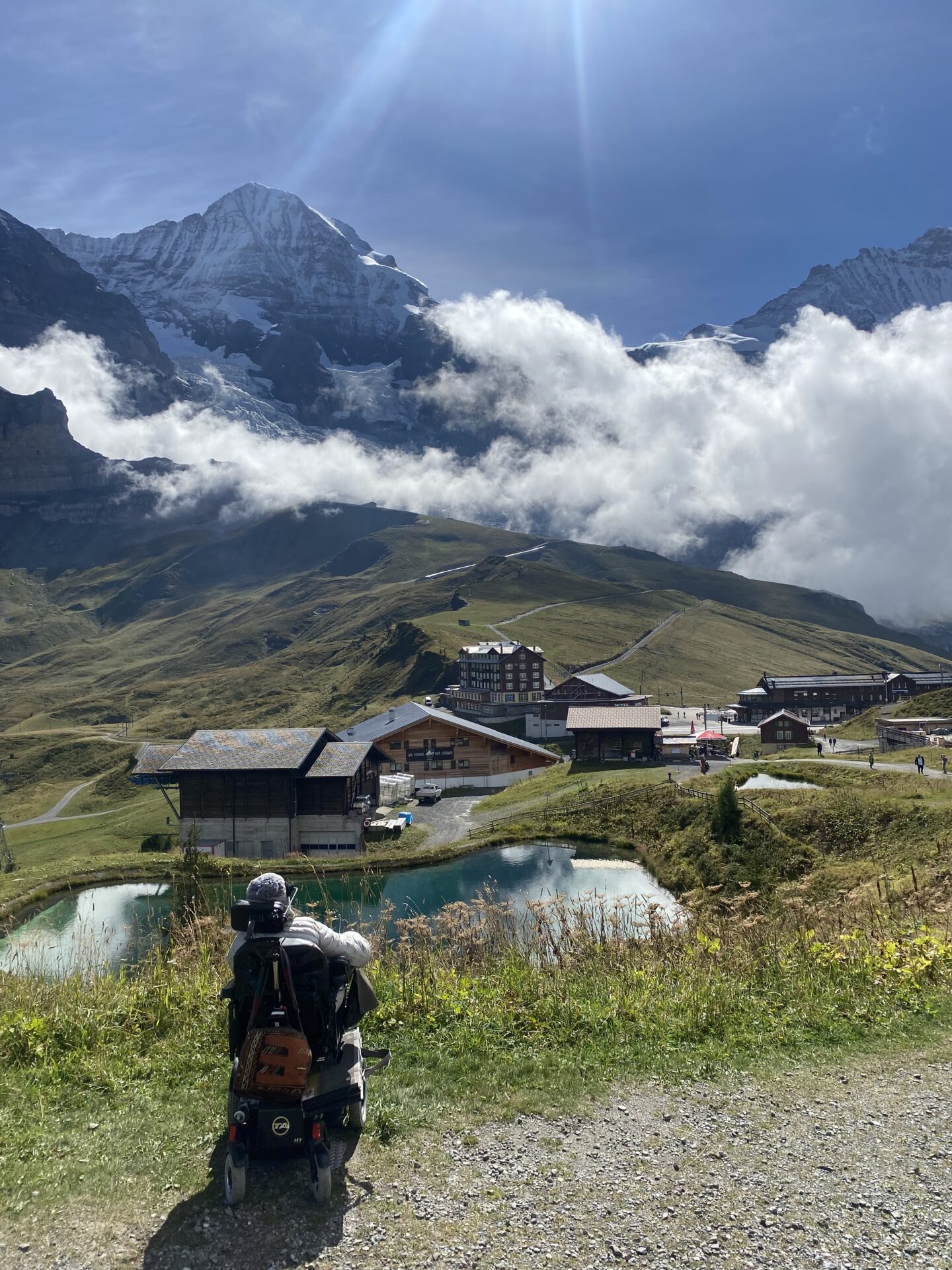 En person i en mobilitetsscooter ser mod alpine bjerge skjult af skyer. I forgrunden er der en dam og flere rustikke bygninger. Solen skinner klart fra toppen af billedet og oplyser scenen med en klar blå himmel.