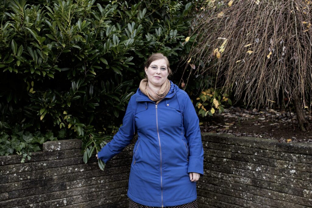 En kvinde i blå frakke står udendørs, lænet op ad en lav stenmur med grønt løv og bare grene bag sig. Hun har et lille smil og bærer briller og et solbrunt tørklæde. Indstillingen er overskyet.