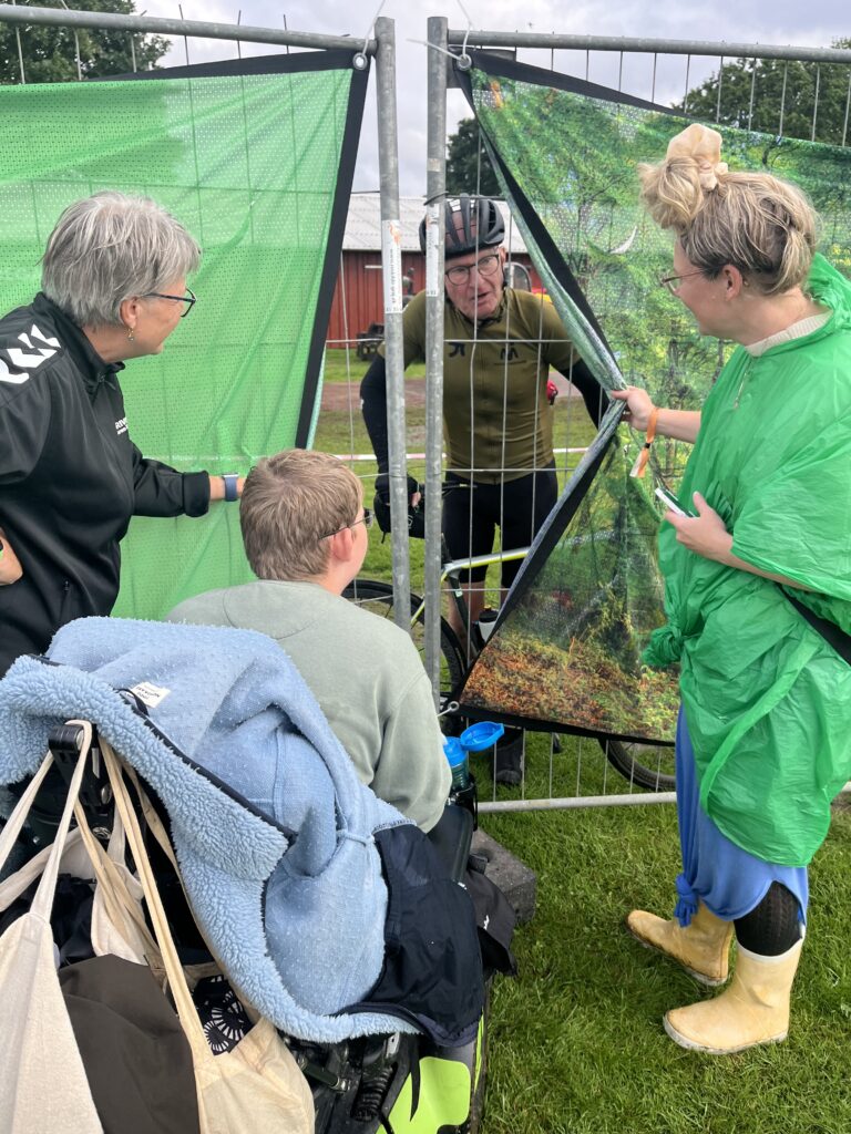 En cyklist holder pause ved et hegn, hvor tre personer hilser på ham. En person i kørestol holder et tæppe, mens de andre stående engagerer sig muntert. Grønt hegn omkranser området, med træer synlige i baggrunden.