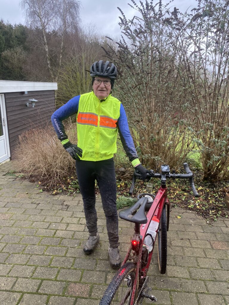 En cyklist iført en lys gul og orange refleksvest står ved siden af en rød cykel på en terrasse. De er klædt i mørke leggings, blå ærmer og en hjelm. Baggrunden har bladløse buske og træer og en overskyet himmel.